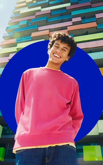 Ein junger Mann in einem rosa Pullover steht vor einem Hochhaus und lacht in die Kamera.