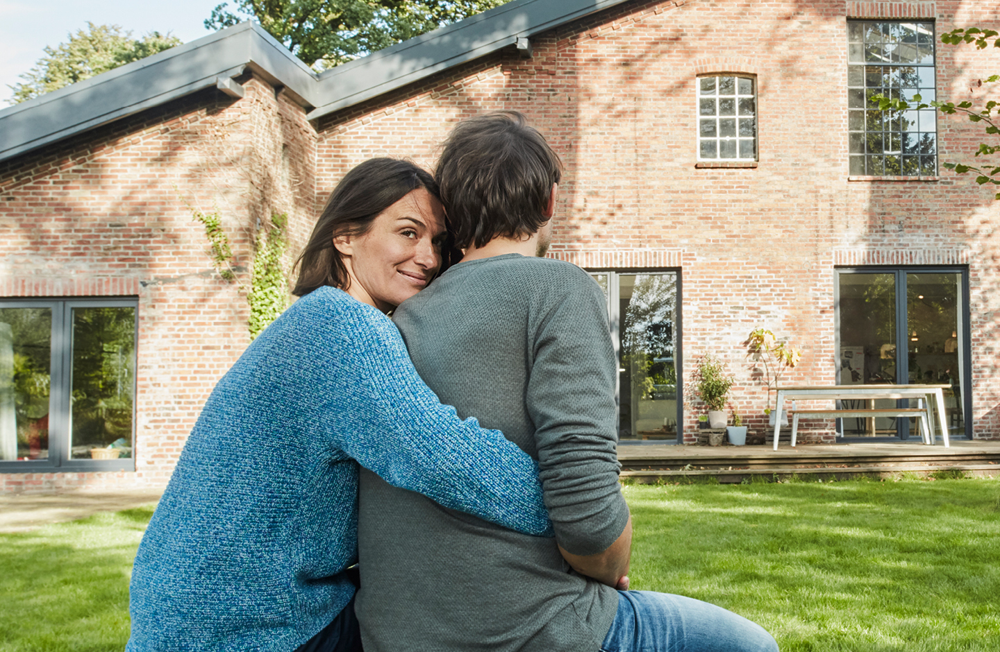 Ein Mann und eine Frau sitzen mit dem Rücken zur Kamera vor einem Haus. Die Frau umarmt den Mann von hinten. Sie blickt über die Schulter in die Kamera.