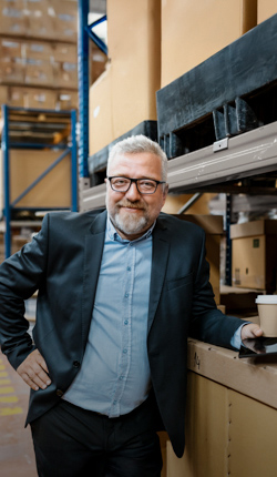 Einb Unternehmer steht in einer Lagerhalle. Er lehnt an einer Kiste und hat ein Tablet und einen Kaffeebecher aus Papier in der Hand. 