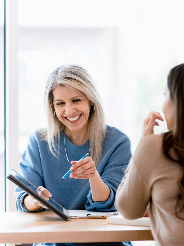 Eine Frau berät eine andere Frau alchend mit Tablet in der Hand und zeigt darauf.