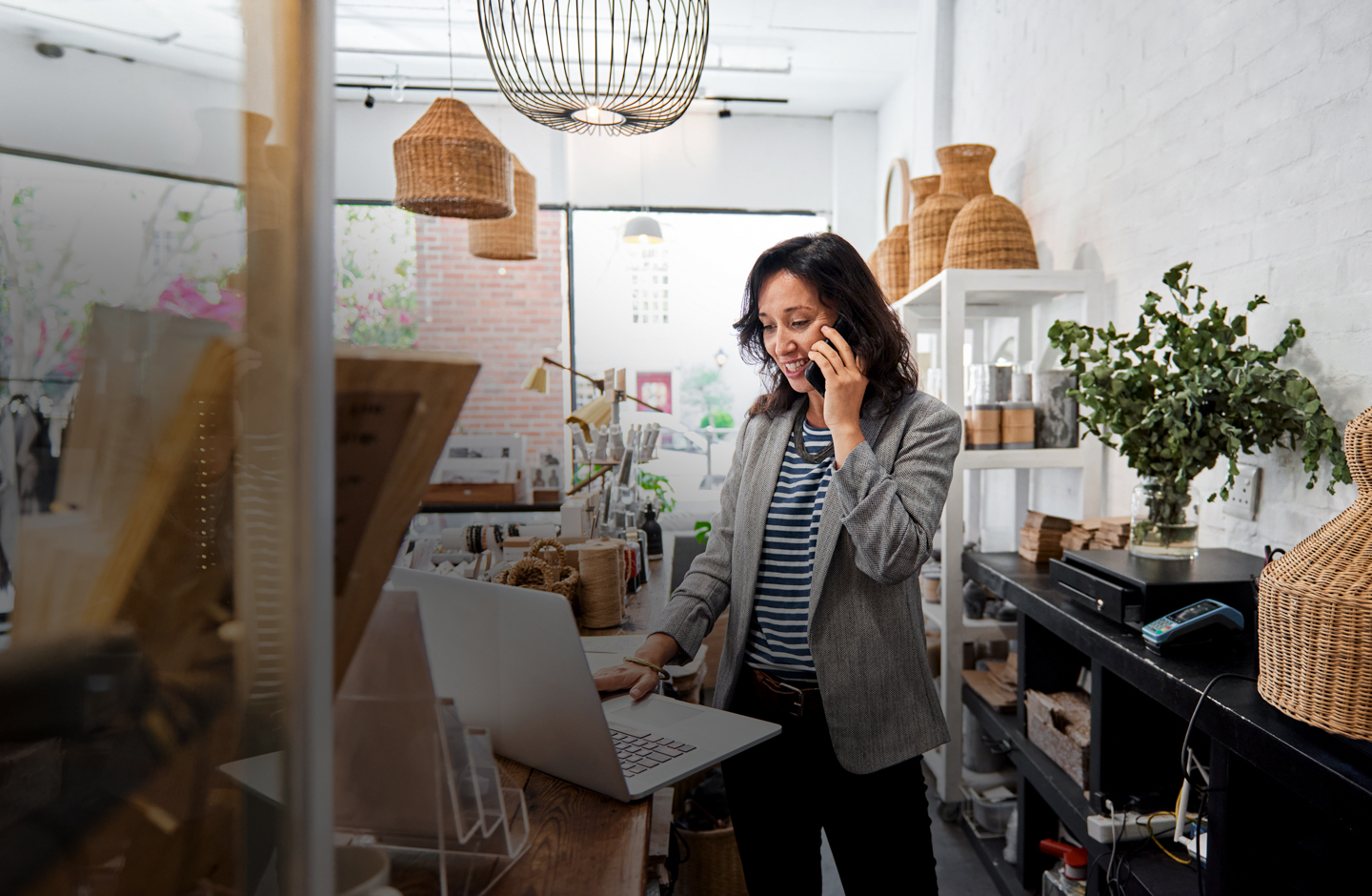 Eine junge Frau steht in einem Geschäft für Korbmaterial-Produkte, telefoniert und blickt dabei auf ihren Laptop.