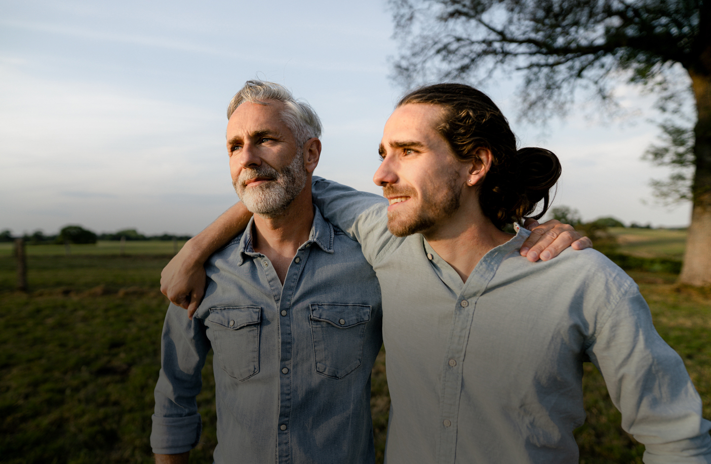 Zwei Männer stehen draußen im Grünen und haben die Arme über die Schulter des jeweils anderen gelegt. Sie blicken lächelnd in die Ferne.